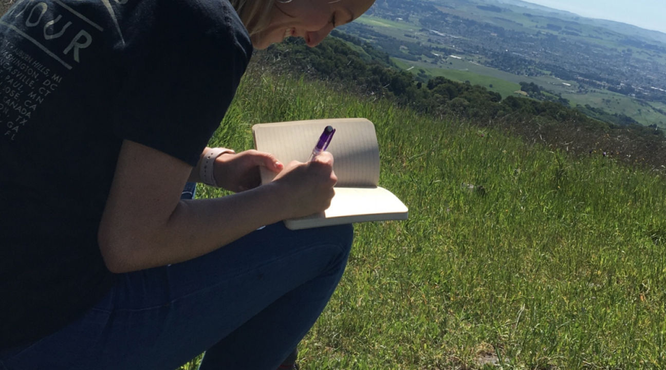 Student sitting in grassy field writing in notebook