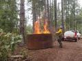 a person stands next to fire in metal kiln in a forest