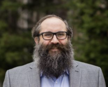 man with beard and glasses smiles in front of trees