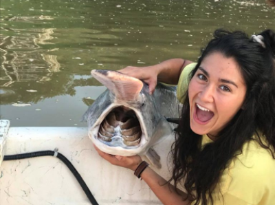 an undergraduate holds a paddlefish on a boat and opens her mouth like it does